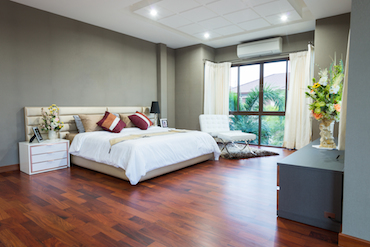 a large room with a hardwood floor, a tall window overlooking trees, a two-person bed with a single bedside table, and a padded chair next to the window.